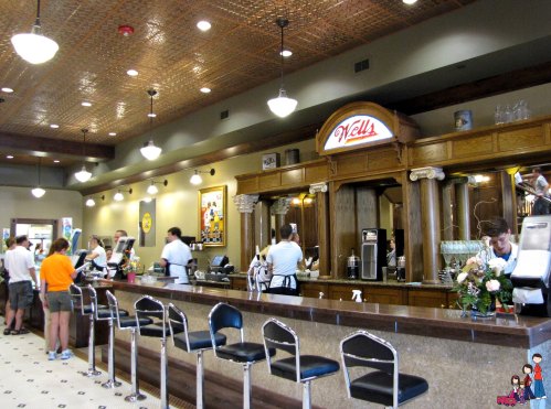 A vintage-style diner interior with a long counter, bar stools, and staff serving customers. Brightly lit and welcoming.