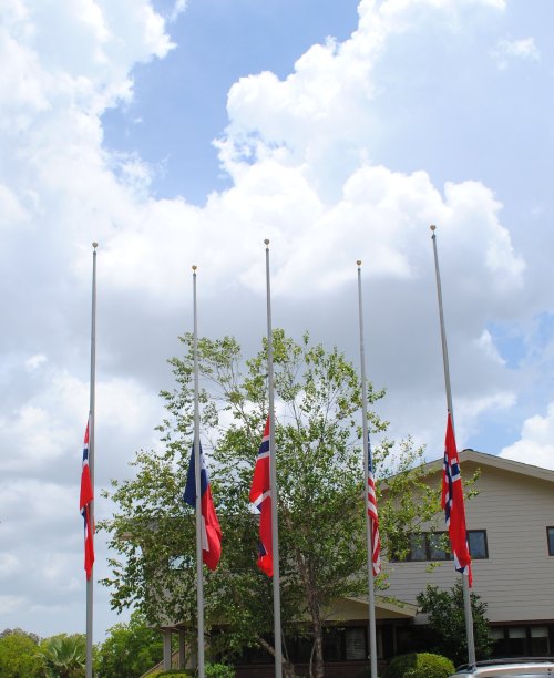 Five flags at half-mast in front of a building, with a partly cloudy sky in the background.