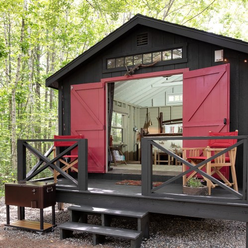 A cozy black cabin with red doors, surrounded by trees, featuring a welcoming porch and rustic interior.