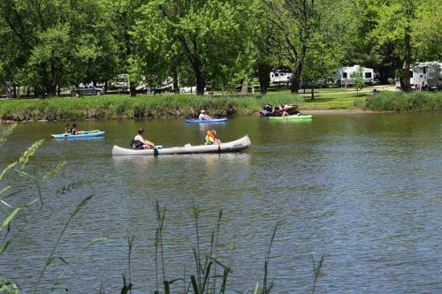 People kayaking on a calm lake surrounded by greenery and trees, with campers visible in the background.
