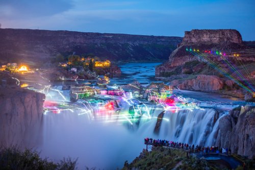 A vibrant waterfall illuminated by colorful lights, with a crowd viewing from a nearby overlook at dusk.