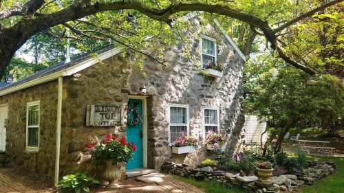 A charming stone cottage with a blue door, surrounded by greenery and colorful flower boxes.