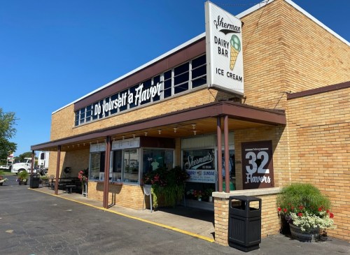 Exterior of Sherman's Dairy Bar, featuring a sign for ice cream and "32 Flavors," with potted plants and a sunny sky.