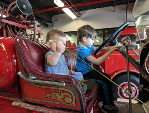 Two young children sit in a vintage fire truck, one holding a toy while the other pretends to drive.