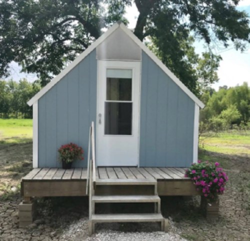 A small, blue and white cabin with a front porch, surrounded by greenery and flower pots.