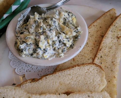 A bowl of creamy spinach dip with herbs, accompanied by slices of toasted bread on a plate.