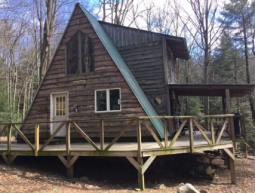 A rustic A-frame cabin surrounded by trees, featuring a wooden deck and a sloped roof.