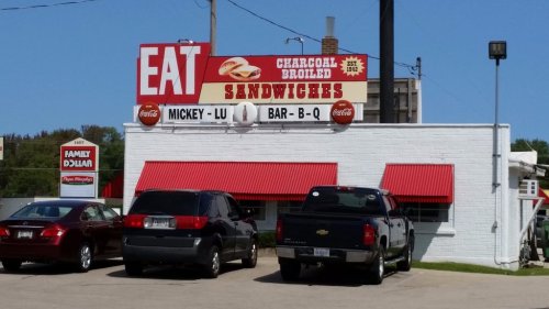 Sign for Mickey-Lu Bar-B-Q featuring "Charcoal Broiled Sandwiches" with red awnings and parked cars in front.