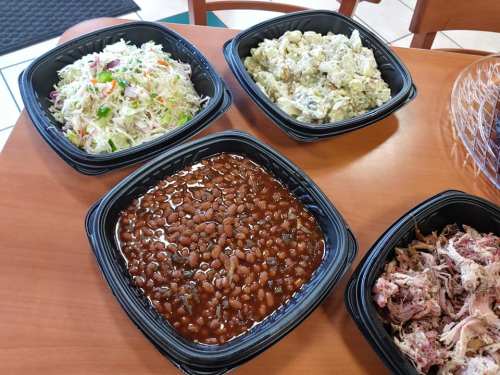 Four containers of food on a table: coleslaw, potato salad, baked beans, and pulled pork.