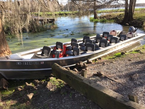 A metal boat with several seats is docked by a grassy area near a calm waterway surrounded by trees.