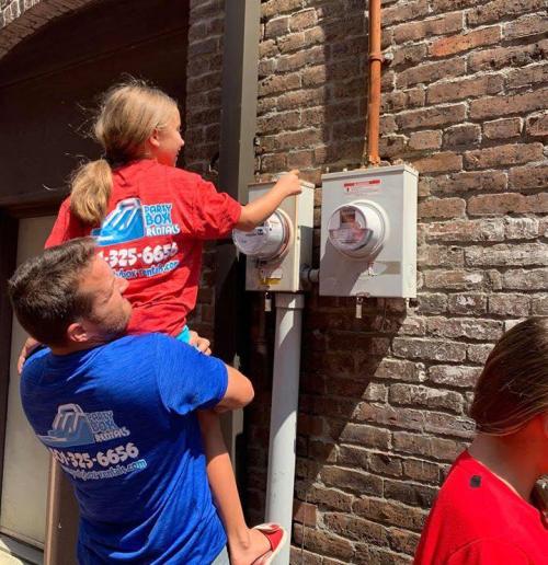A man lifts a girl to reach electric meters on a brick wall, while another person stands nearby.