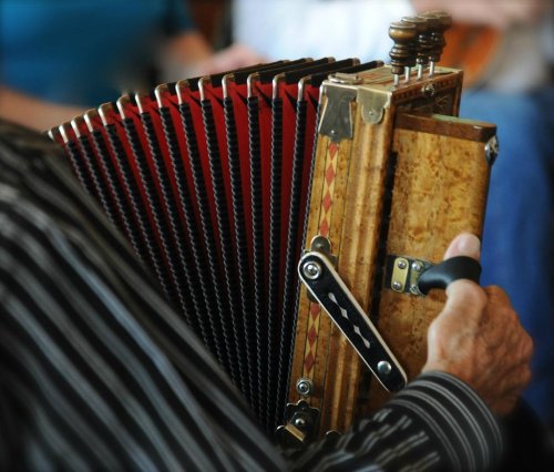 A close-up of a person's hand holding a wooden accordion with red and black bellows, set against a blurred background.