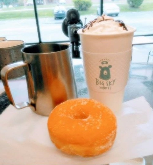 A glazed donut on a napkin next to a metal cup and a coffee cup from Big Sky Donuts.