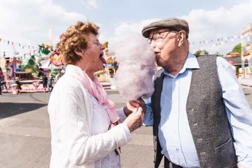 An elderly couple playfully sharing cotton candy at a fair, smiling and enjoying each other's company.