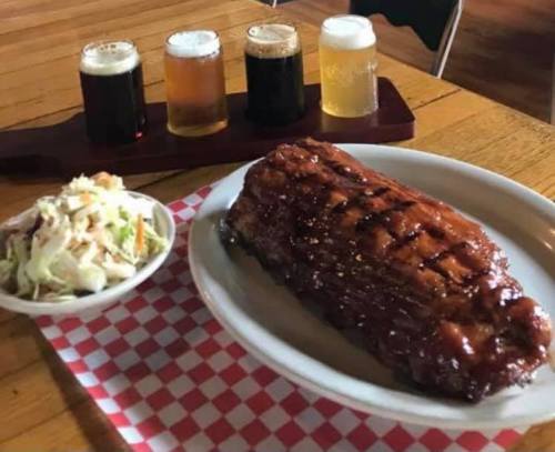A plate of glazed ribs with coleslaw, accompanied by a flight of four craft beers on a wooden tray.
