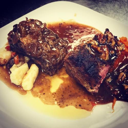 A plate featuring two pieces of meat with a rich sauce, served with gnocchi and a colorful vegetable medley.