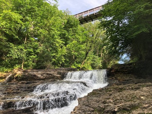A serene waterfall cascades over rocks, surrounded by lush green trees, with a wooden bridge above.