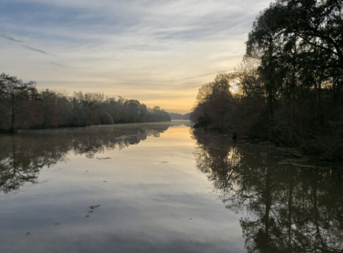 A serene river at sunset, reflecting trees and a colorful sky, with calm waters and a peaceful atmosphere.