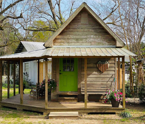 A charming wooden cottage with a green door, surrounded by flowers and a porch, set in a serene outdoor environment.