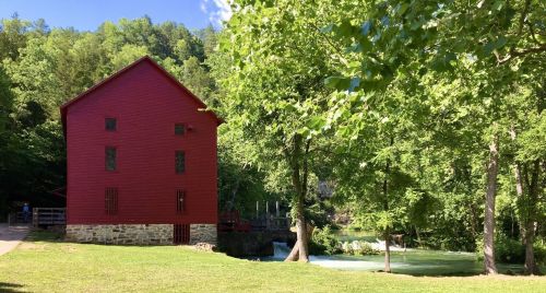 A red barn-style building beside a river, surrounded by lush green trees and grass under a clear blue sky.