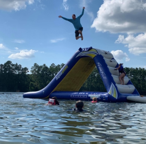 A person jumps off an inflatable water slide into a lake, while others swim nearby under a blue sky.