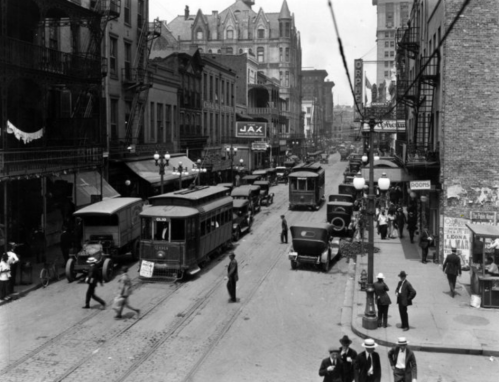 Historic black-and-white street scene with vintage cars, streetcars, and pedestrians in a bustling urban setting.