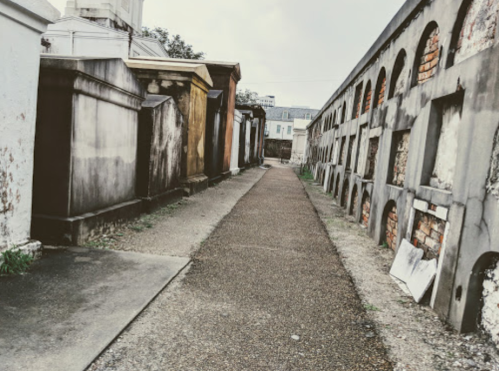 A narrow pathway between old, weathered tombs and mausoleums in a cemetery, with a cloudy sky above.