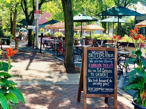 A sidewalk scene with a sign for Setteluna, announcing curbside pickup and a welcoming message, surrounded by greenery.
