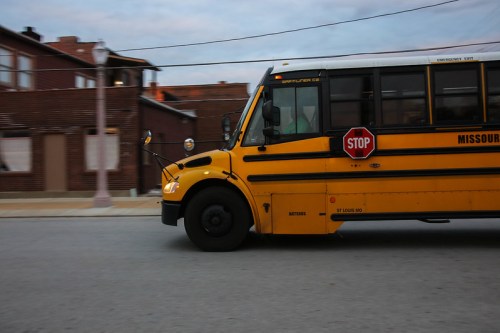 A yellow school bus with a stop sign, driving past a brick building on a cloudy day.