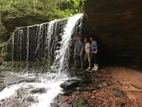 Three people stand under a waterfall in a lush, green forest, with water cascading over rocks.