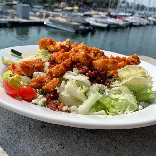 A plate of salad topped with crispy chicken, bacon bits, and cherry tomatoes, with a marina in the background.