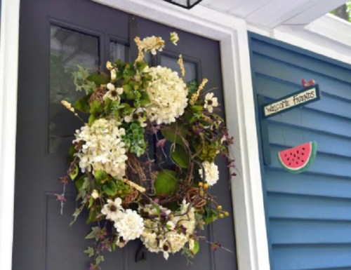 A decorative wreath of white flowers and greenery hangs on a dark door beside a sign that says "Welcome Friends."