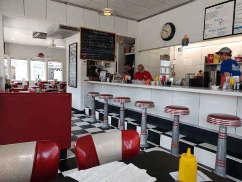 A retro diner interior with red and silver stools, a counter, and a menu board. Staff are visible in the background.