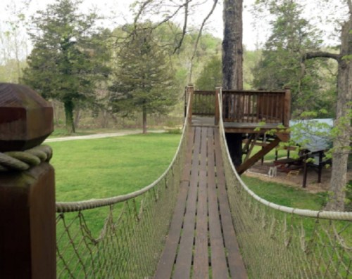 A wooden rope bridge leads to a treehouse, surrounded by greenery and trees in a peaceful outdoor setting.