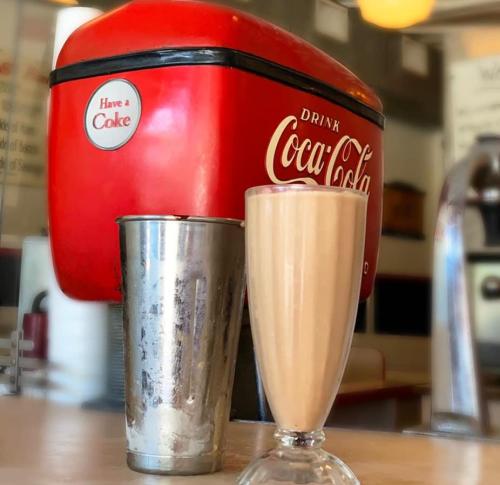 A vintage Coca-Cola dispenser next to a tall glass of creamy milkshake and a metal shaker.