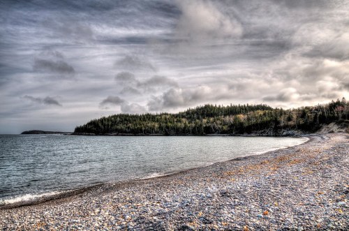 A rocky beach with a calm sea, surrounded by lush green hills under a cloudy sky.