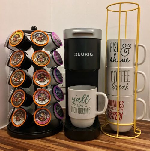 A Keurig coffee maker with a carousel of coffee pods and stacked mugs that say "Y'all have a good morning" and "Rise & shine."