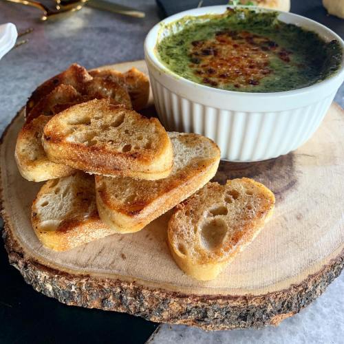 A bowl of green spinach dip sits on a wooden platter, accompanied by toasted bread slices.