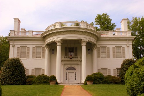 A grand white mansion with a circular portico, tall columns, and manicured lawns surrounded by lush greenery.