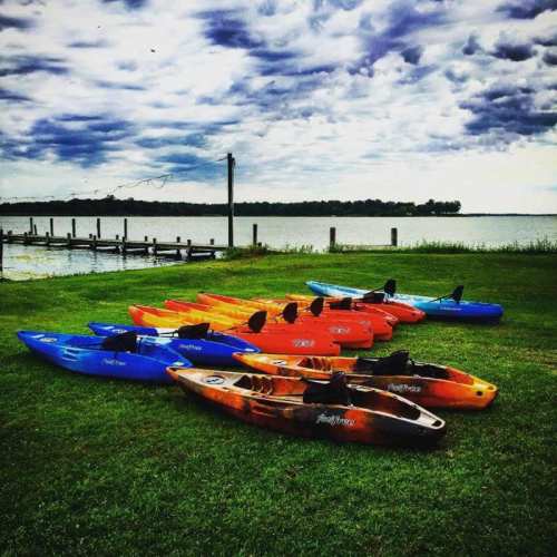 Colorful kayaks lined up on green grass by a calm lake under a cloudy sky.