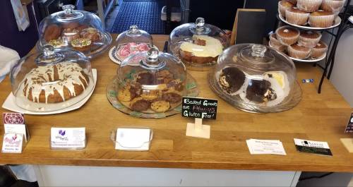 A display of various baked goods under glass domes, including cakes, cookies, and muffins, on a wooden counter.