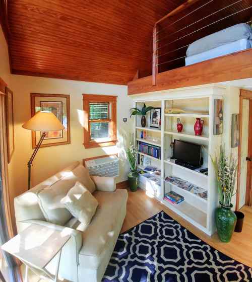 Cozy living space with a sofa, lamp, bookshelf, and a lofted bed, featuring warm wood accents and natural light.