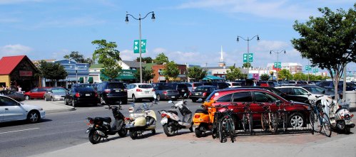 A busy street scene with parked cars, scooters, and bicycles, featuring green banners and a clear blue sky.