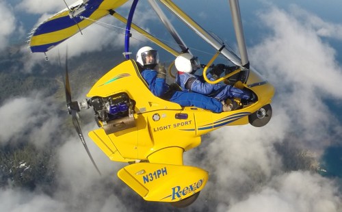 A yellow light sport aircraft flying above the clouds with two pilots inside, enjoying a scenic aerial view.