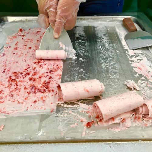 A person in gloves uses a metal spatula to roll pink ice cream on a cold surface, with some rolled ice cream pieces visible.