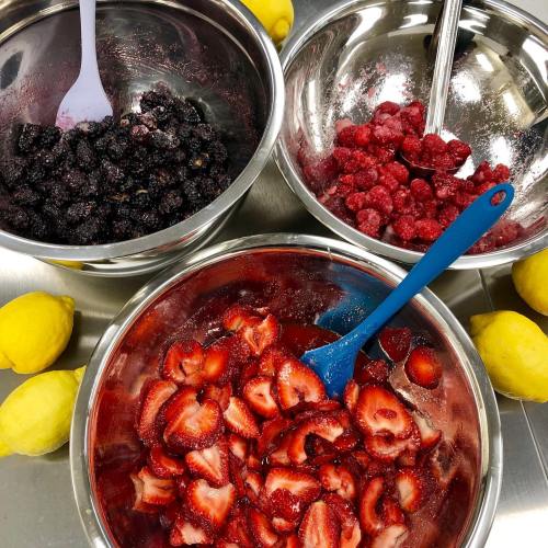 Three metal bowls contain blackberries, raspberries, and sliced strawberries, with lemons scattered around.