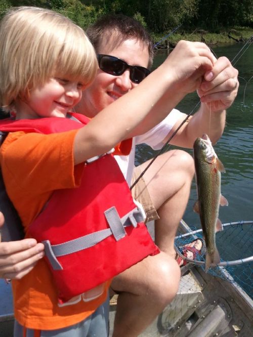 A child in a life jacket proudly holds a fish while an adult smiles beside him on a boat.