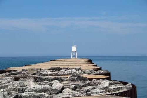 A long wooden pier extends into calm blue waters, leading to a white lighthouse under a clear sky.