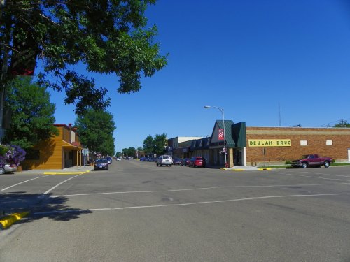 A quiet street scene featuring shops, parked cars, and clear blue skies in a small town.