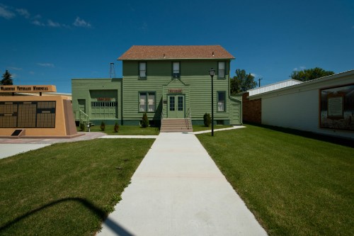 A green two-story building with a brown roof, surrounded by grass and a clear blue sky. Pathway leads to the entrance.